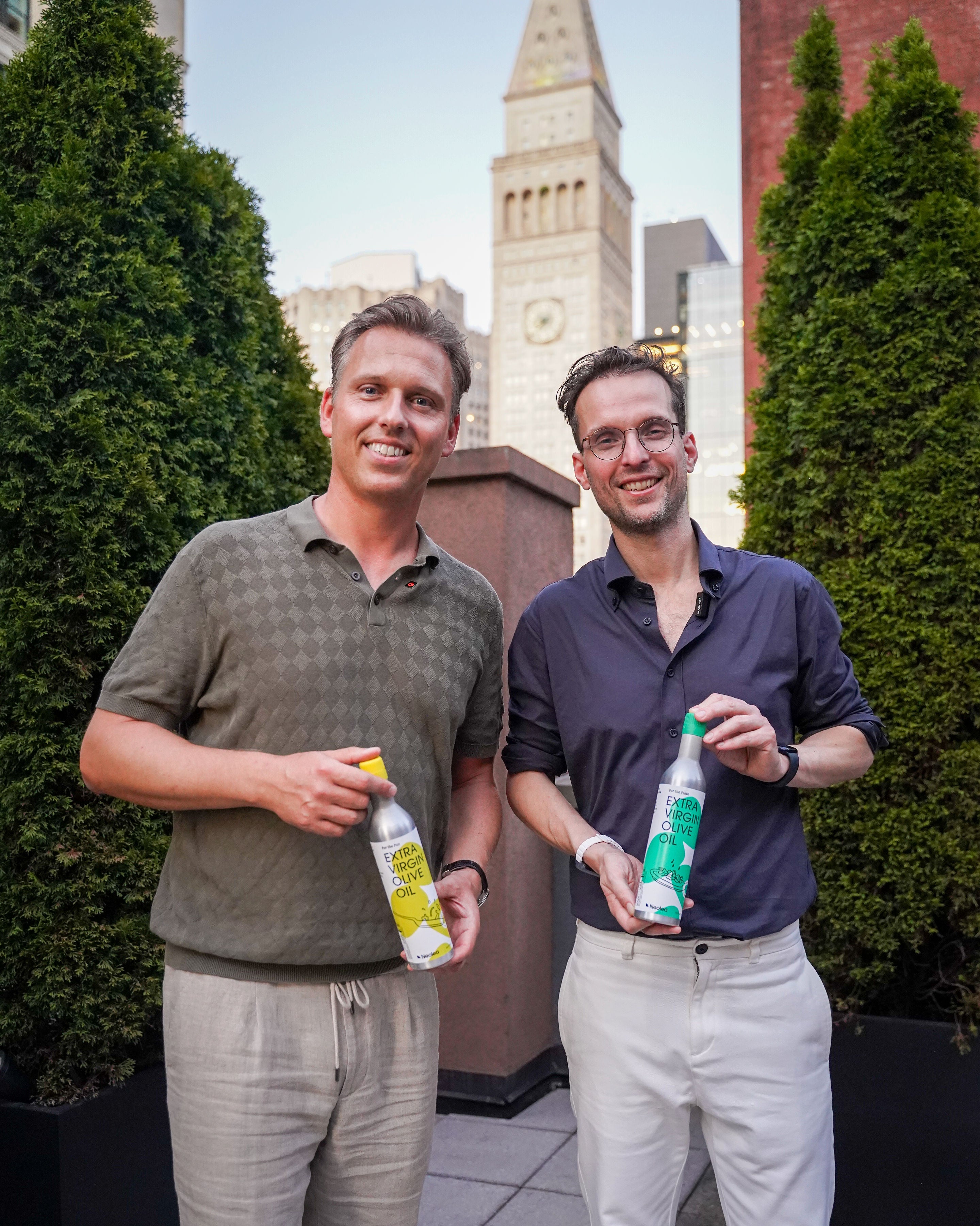 Two men holding bottles outdoors with a cityscape background