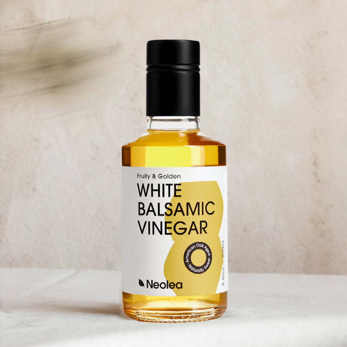 A bottle of Neolea White Balsamic Vinegar with a black cap and white label, set on a light marble surface against a textured beige background.